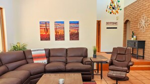 A living room with a brown leather sofa, a coffee table, and a chair. There are three paintings on the wall, a chandelier, and a sign that says "BE OUR GUEST". - Close to downtown but feels like country-Very Private! Refreshing pool & hot tub (Tucson)