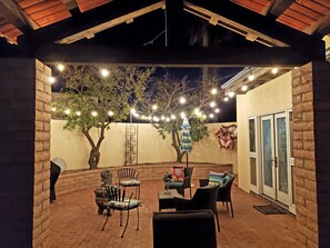 Un patio al aire libre con suelo de ladrillo, una zona de estar cubierta con sillas y mesas, luces colgantes y un árbol.