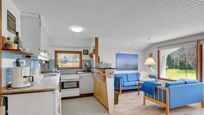 Une cuisine compacte équipée d’une cuisinière, d’un évier et d’une machine à café. Un canapé bleu et une lampe se trouvent dans l’espace salon, avec un tableau accroché au-dessus.