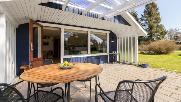 Une terrasse couverte avec une table et des chaises en bois, un saladier rempli de fruits et une vue sur l’intérieur de la maison à travers de grandes fenêtres.