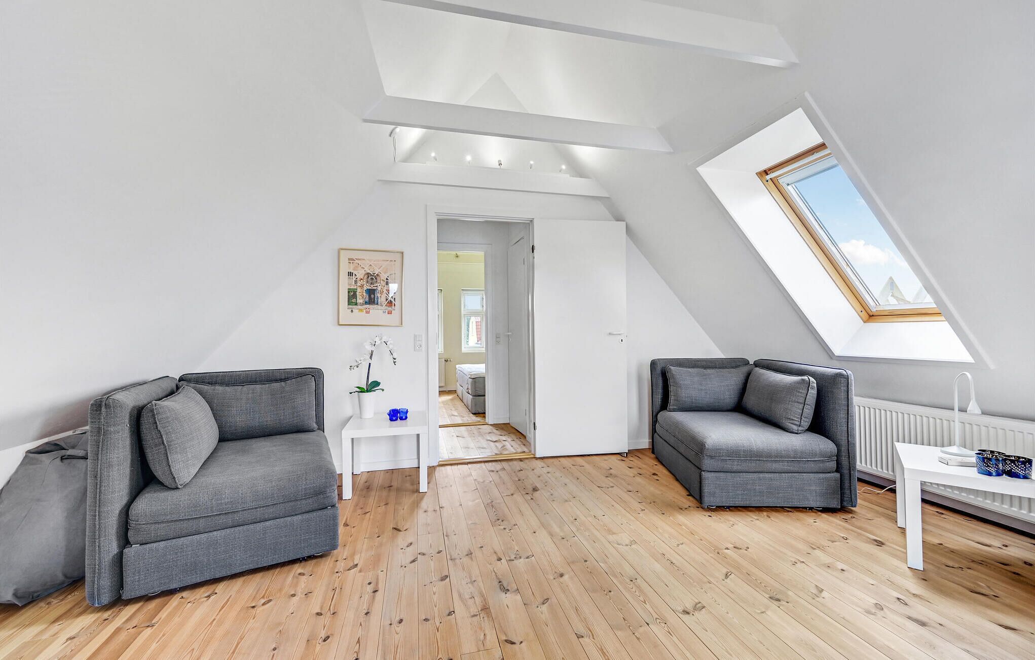 A modern loft apartment with a sloped ceiling, wooden flooring, two grey sofas, a white side table, and a skylight.