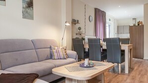 A modern living room with a grey sofa, a small wooden table, and a dining area with chairs and a kitchen in the background. - 5 bedroom amazing home in Wurster Nordseeküste (Wurster Nordseeküste)