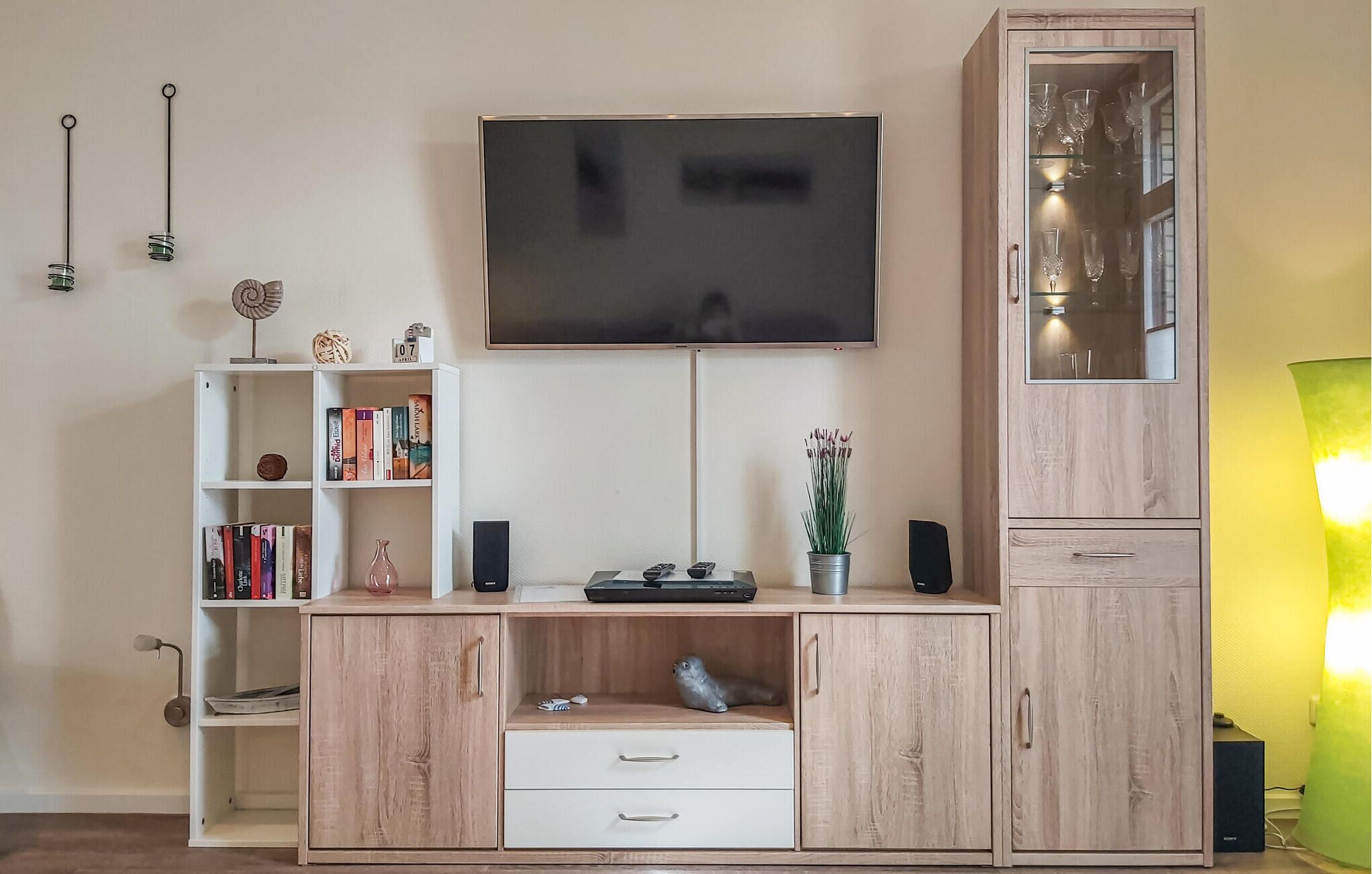 A living room with a TV mounted on the wall, a bookshelf, a cabinet with glass doors, and a lamp.