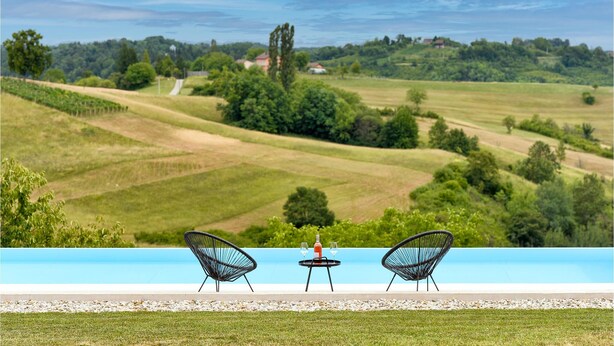 Vista de la piscina con dos sillas y una mesa, con vistas a un paisaje exuberante con campos y árboles.
