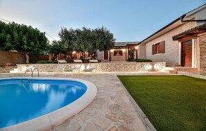 A backyard with a swimming pool, stone patio, and a grassy lawn.