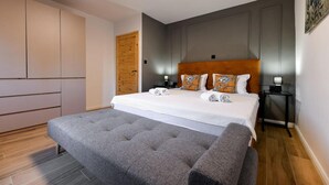 A modern bedroom with a large bed, a grey upholstered bench, a wooden door, and a nightstand with a lamp. - Nice home in Lisicic with sauna (Lisicic)