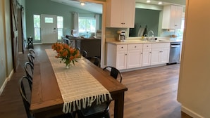 A dining area with a wooden table, chairs, and a vase of flowers. There is a kitchen in the background with white cabinets and a dishwasher. - A charming country escape.  Near wineries, SLO, hiking, and SM Ranch & lake. (Santa Margarita)