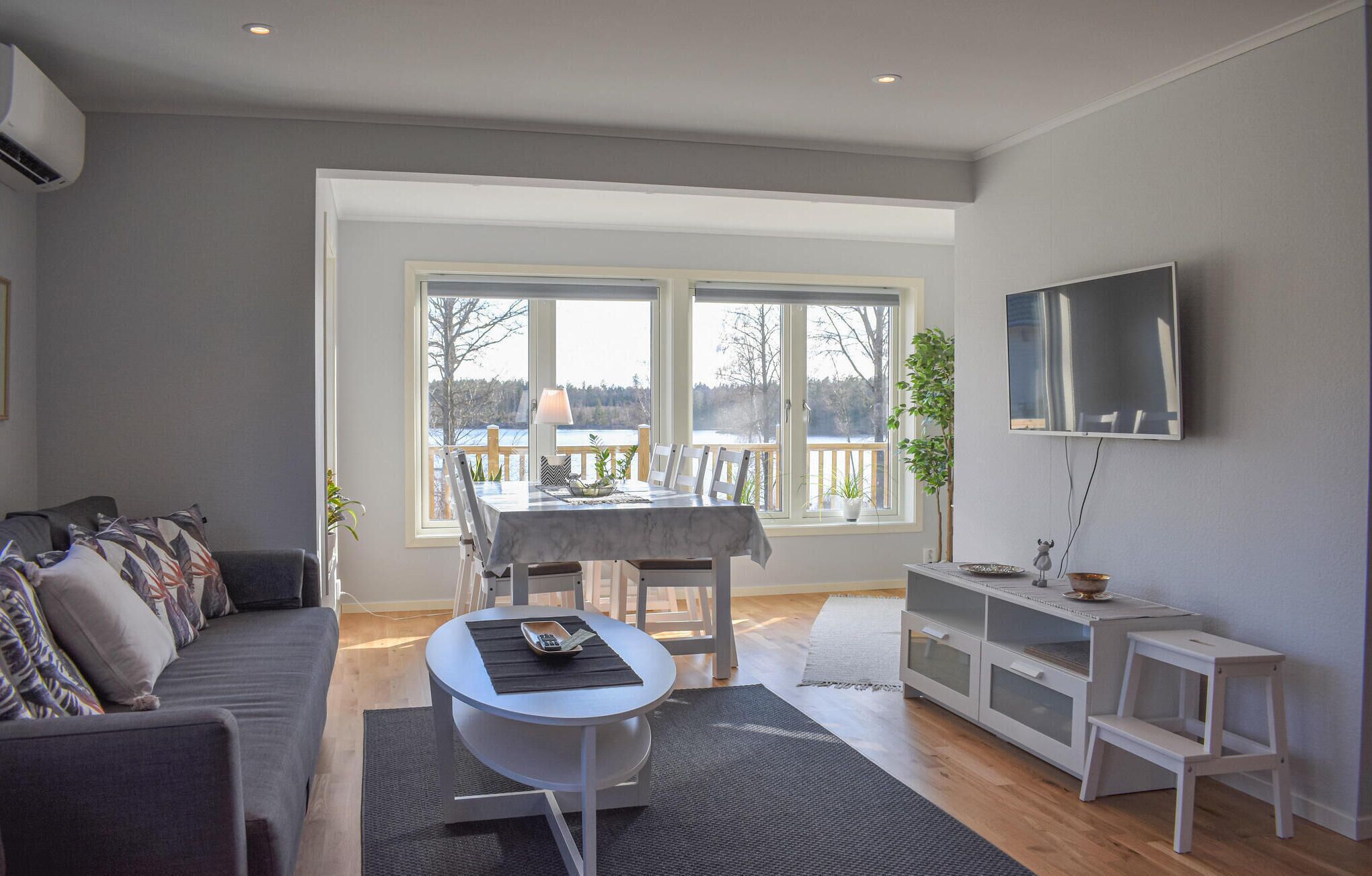 Ein Wohnzimmer mit einer Couch, einem Esstisch mit Stühlen, einem an der Wand befestigten Fernseher und Blick ins Freie durch große Fenster.