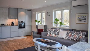 A modern kitchen with grey cabinets and a built-in oven, a grey sofa with patterned cushions, a coffee table, and a window with a view of trees.