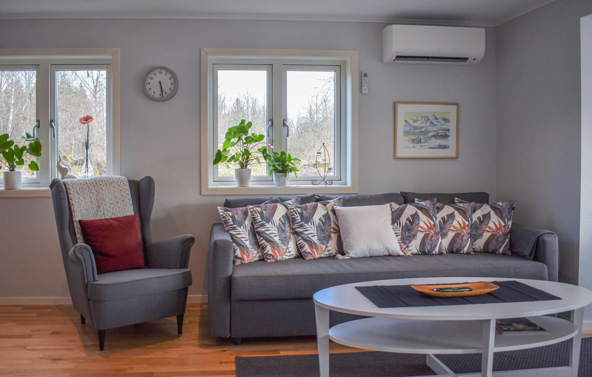 A living room with a grey sofa, a grey armchair, a round white table, and two windows with potted plants.