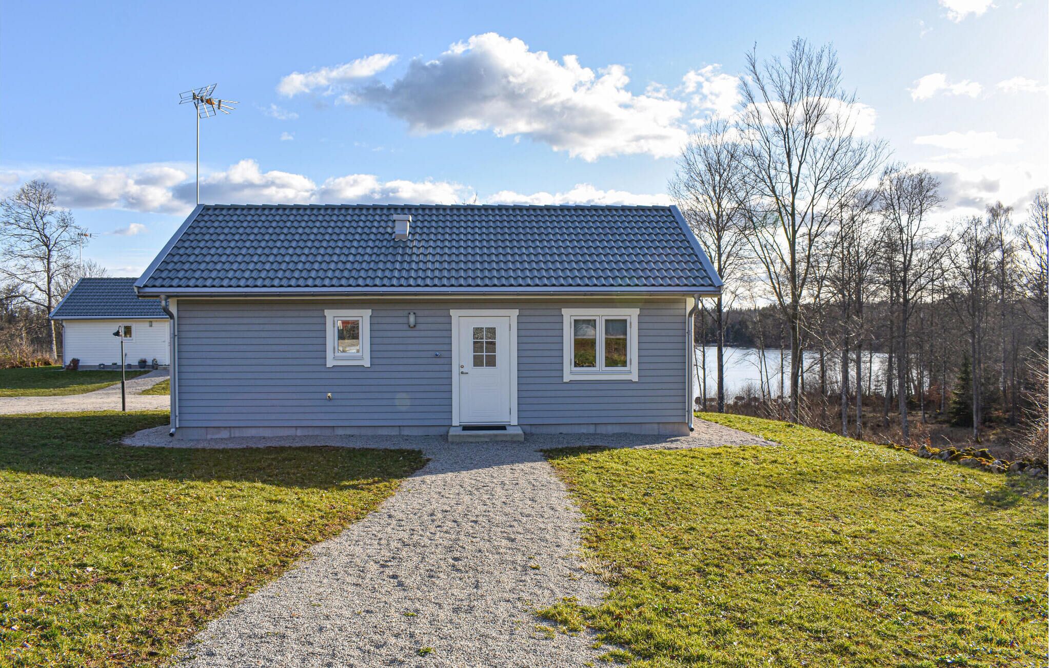 A small, single-story house with a blue roof and a white door, located near a body of water with trees in the background.