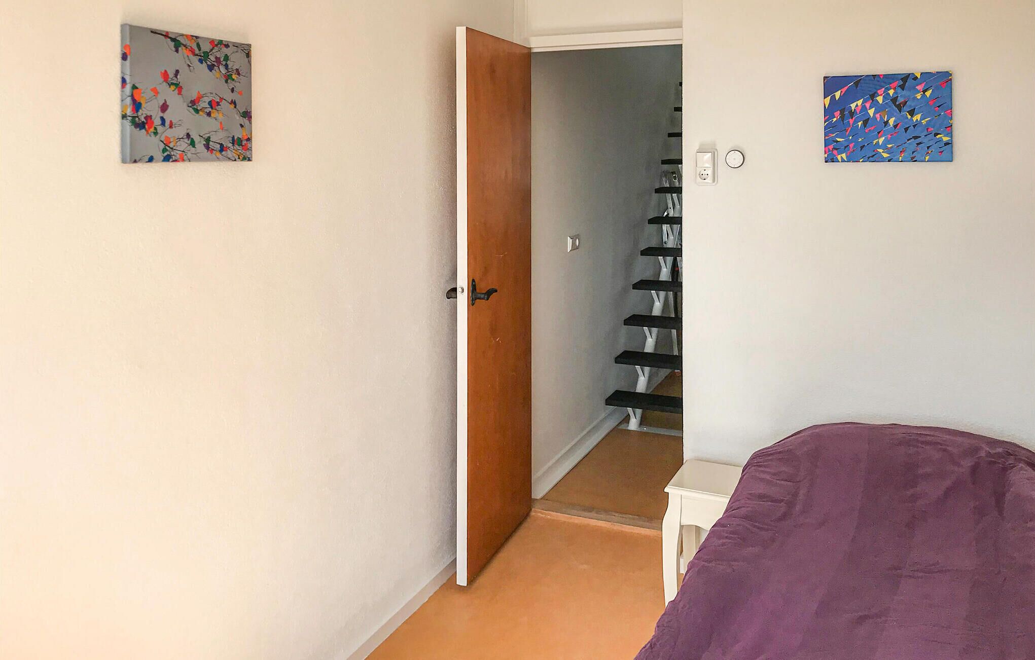 Una habitación con una cama morada, una puerta de madera y una escalera que lleva a otra habitación. Hay dos cuadros en las paredes.