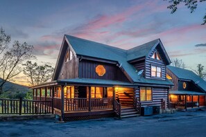 Hirsimökki, jossa on kuisti ja useita ikkunoita, ja taustalla auringonlaskun värittämä taivas.