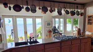 A kitchen with a large island, wooden cabinets, and hanging pots. There is a view of a body of water through large windows. - Beachfront Home With Sweeping Views of Water and Mount Baker (Port Ludlow)