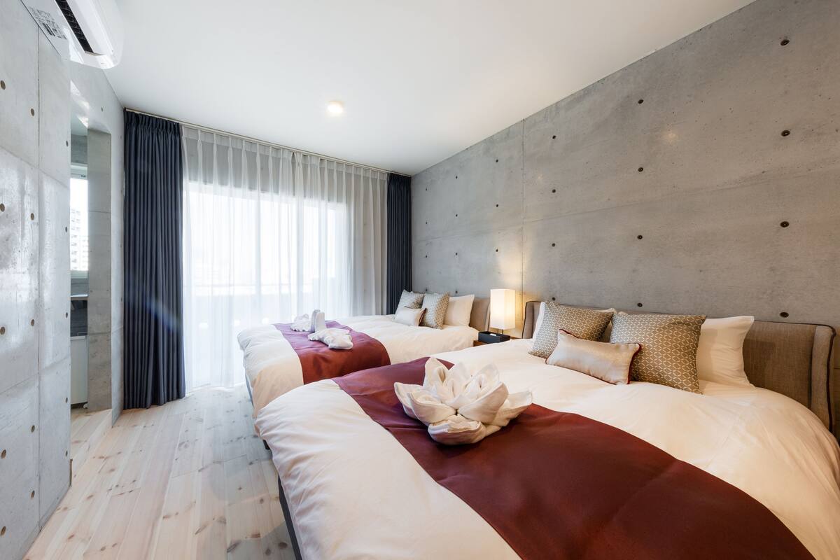 Habitación de hotel con dos camas, un ventanal con cortinas, una pared de detalles de concreto y piso de madera.