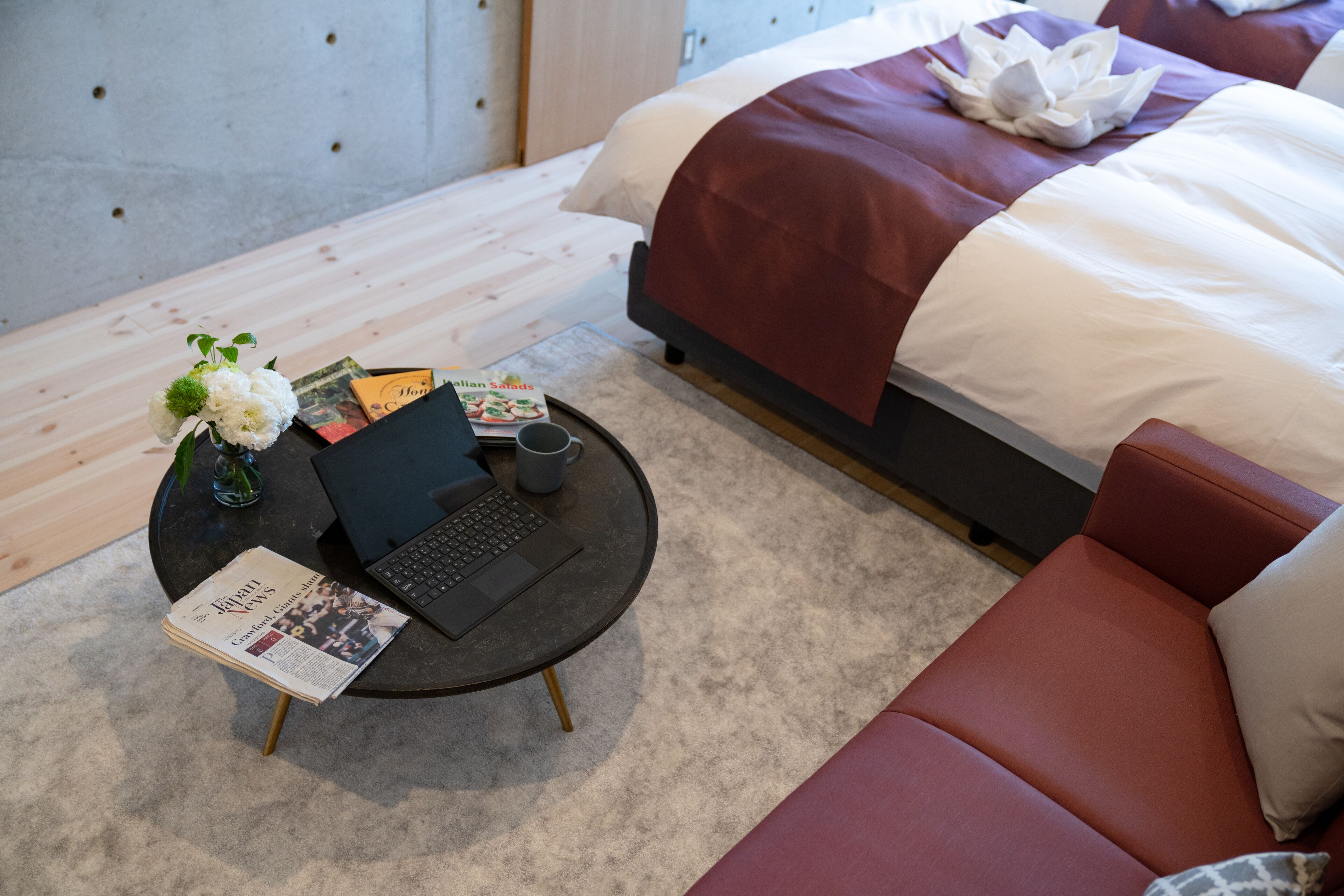 Una habitación de hotel ordenada con una cama, una mesa de centro redonda con una computadora portátil, un jarrón con flores y una revista en el suelo.