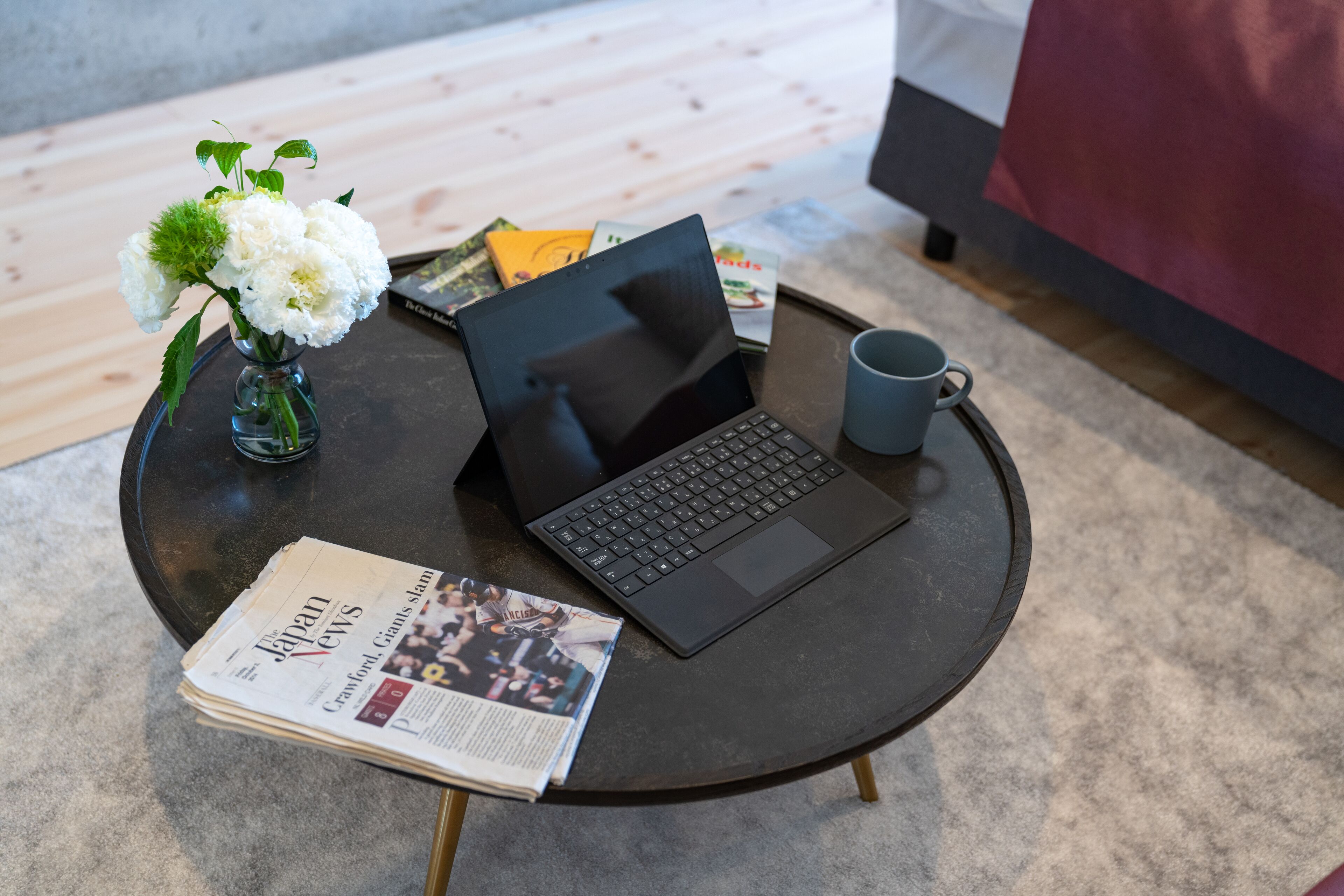A laptop on a round table with a newspaper, a mug, and a vase with flowers.