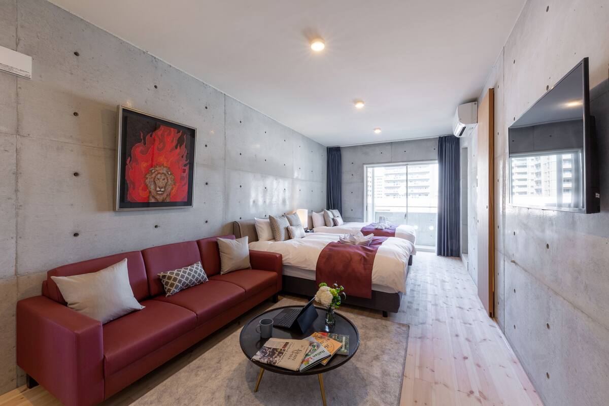 Una habitación de hotel moderna con un sofá rojo, una cama grande, una mesita de centro con libros y un cuadro en la pared.