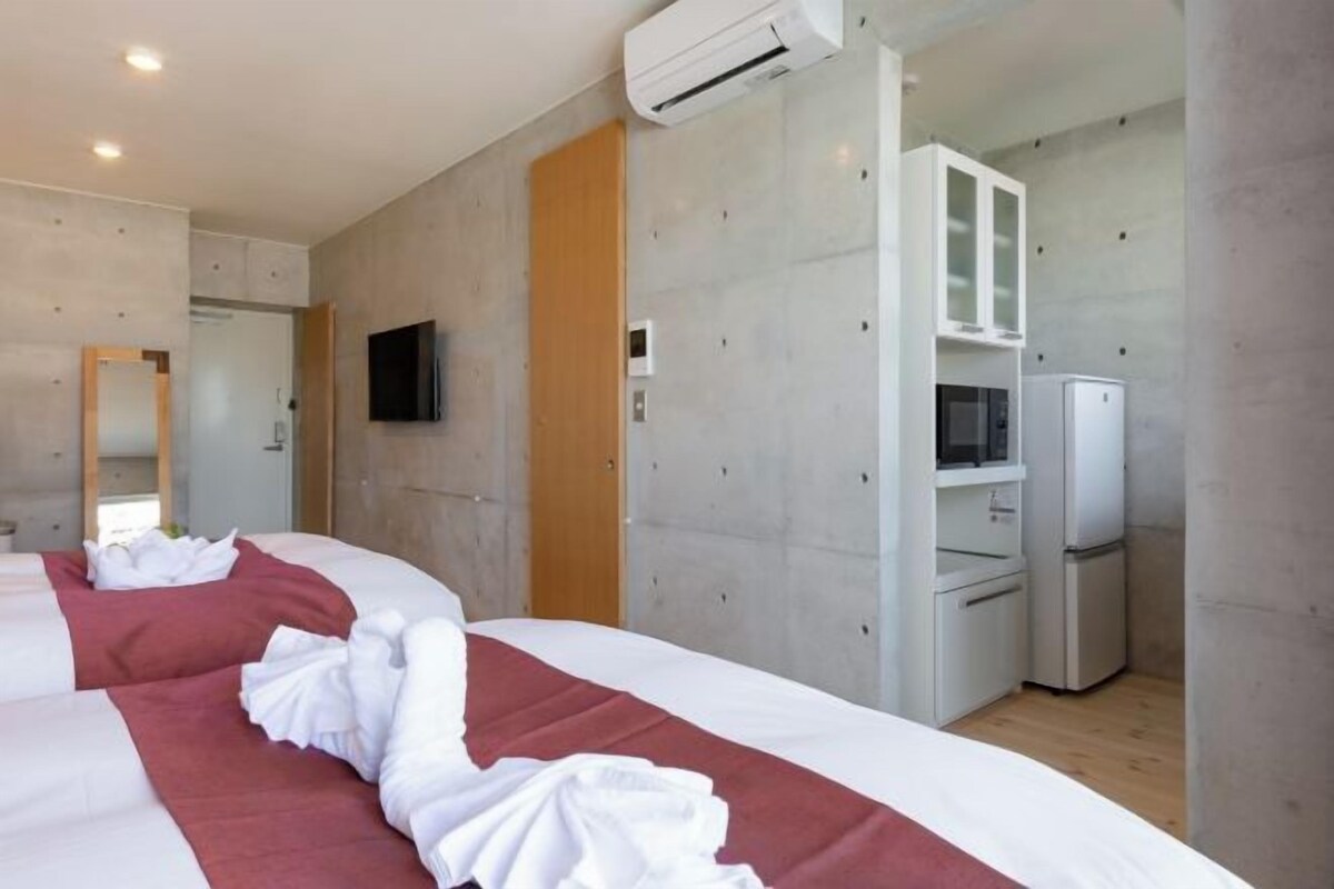 A modern hotel room with a large bed, a flat-screen TV mounted on the wall, a built-in wardrobe, and a kitchenette area with a microwave and refrigerator.
