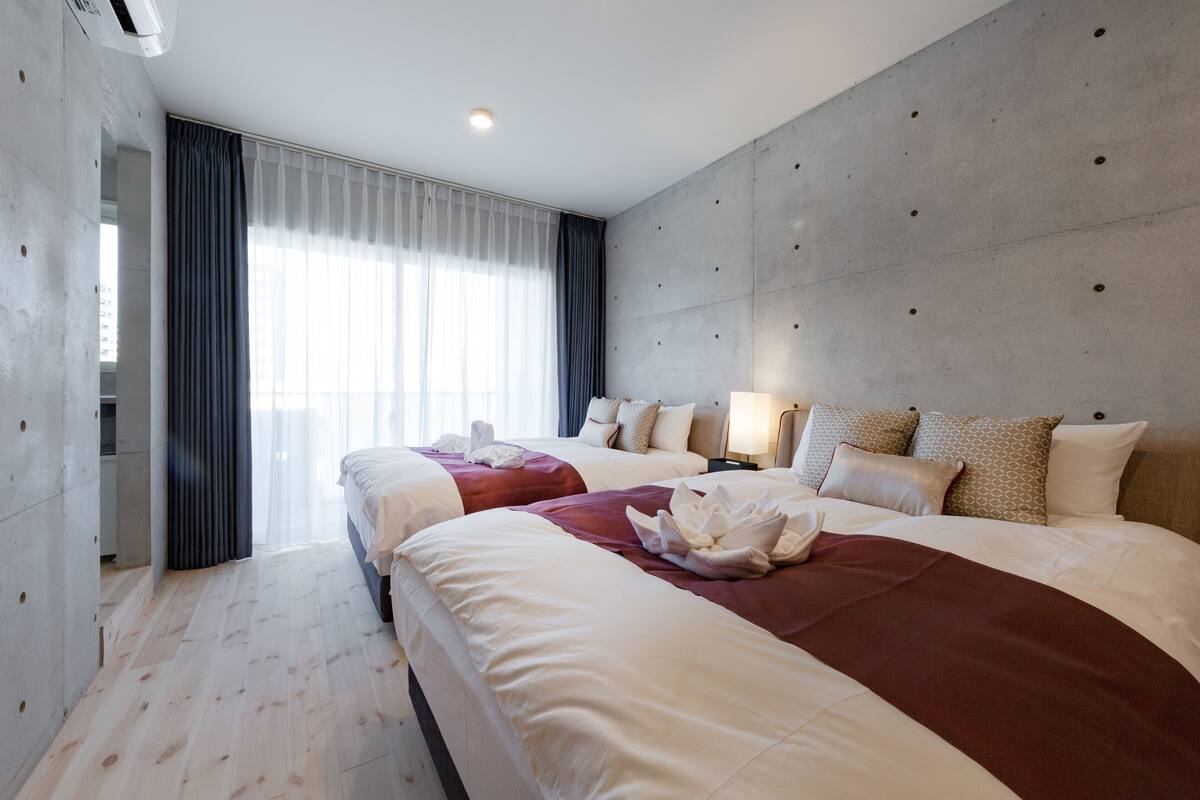 Habitación de hotel con dos camas, una pared de acento de hormigón, un ventanal con cortinas y suelo de madera.