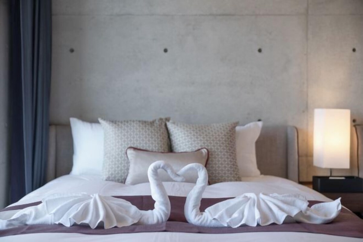 A hotel room with a bed, pillows, and a nightstand with a lamp.