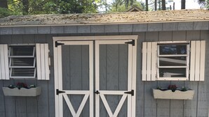 A small, single-story building with a gray exterior and white trim. It features a large, double door with a cross-brace design, flanked by two windows with shutters. There are two flower boxes on the exterior walls, each containing red flowers.