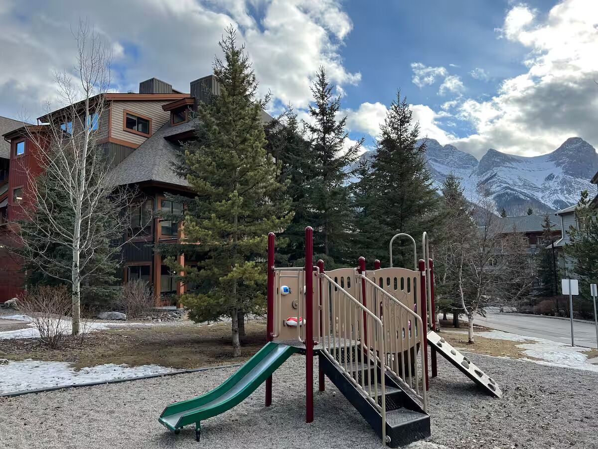 A playground with a slide and climbing structures, surrounded by trees and buildings, with mountains in the background.