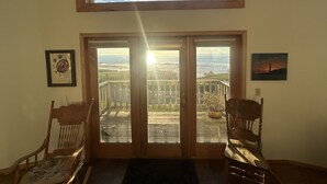 A room with a large window overlooking a coastal area, including a bridge and ocean. There are two wooden chairs and a framed picture on the wall.