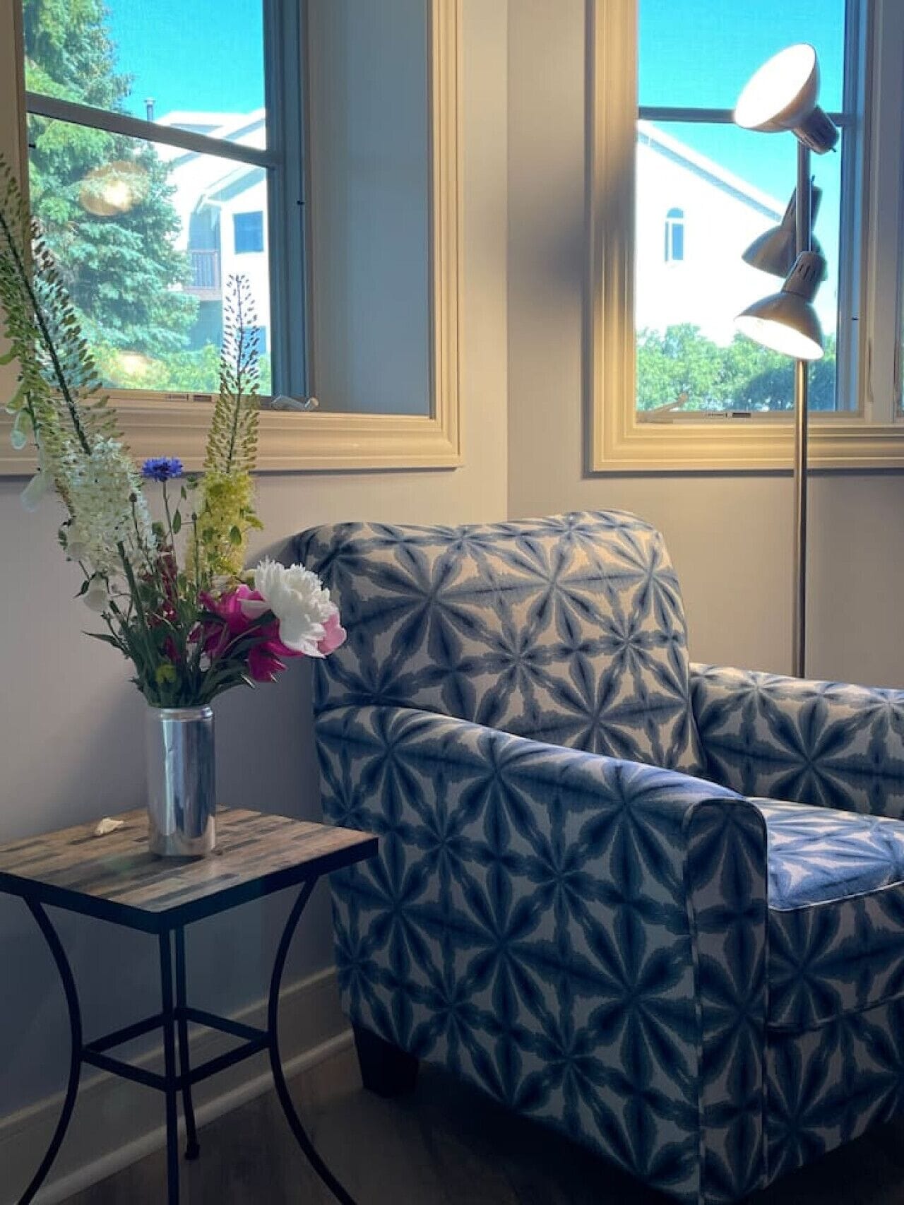 Una habitación con un sillón estampado, una mesita con un jarrón de flores y una lámpara de pie.