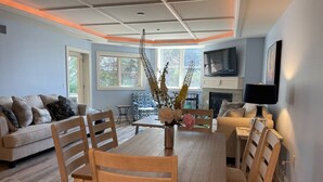 A dining area with a wooden table and chairs, a vase with flowers, and a television in the background. - Beachy Condo with Pool & Hot Tub (Traverse City)