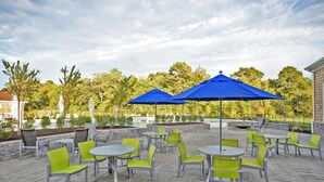 An outdoor patio with round tables and chairs, blue umbrellas, and a pool in the background. - Bright & Airy House. Child friendly, EV charging, Pool, sauna, gym, kayak+ (Ocean View)