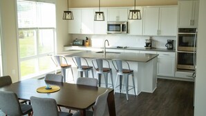 A modern kitchen with white cabinets, a central island with bar stools, a dining table with chairs, and pendant lighting. - Bright & Airy House. Child friendly, EV charging, Pool, sauna, gym, kayak+ (Ocean View)