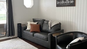 Ein Wohnzimmer mit grauem Sofa, zwei schwarzen Ledersesseln, einem weißen Teppich und einem Fenster mit Blick auf Grünflächen.
