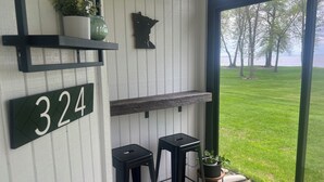 Eine Veranda mit einem Hinweisschild, zwei Barhocker, einem Regal mit einer Pflanze und Blick auf eine Rasenfläche.
