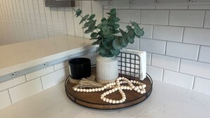 Eine Küchenarbeitsplatte mit weiß gefliestem Fliesenspiegel, einer schwarzen Tasse, einer weißen Vase mit grünen Blättern und einem Holztablett mit einer Reihe weißer Perlen.