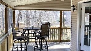 A covered outdoor deck with a table and chairs, a wooden railing, and a view of trees. - Charming Riverside Cottage on 5 Acres- Perfect for Couples & Nature Lovers (Amherst)
