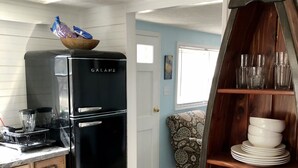 A kitchen with a black Galanz refrigerator, wooden cabinets, a patterned rug, and a wooden shelf with dishes and jars. - Charming Riverside Cottage on 5 Acres- Perfect for Couples & Nature Lovers (Amherst)