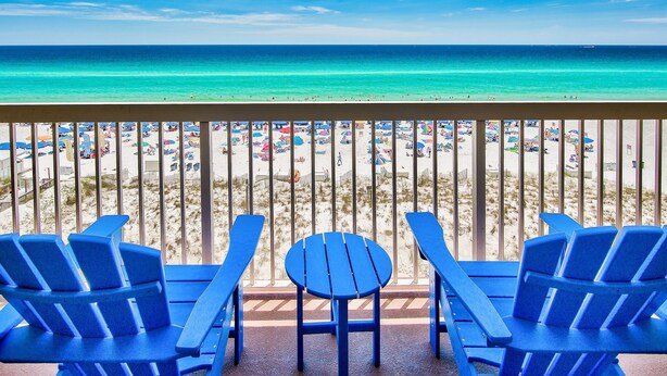 A balcony with blue chairs and a table overlooking a beach with umbrellas and people. - Renovated beachfront condo, spectacular Gulf views (Destin)