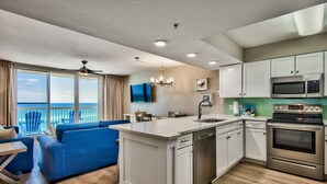 A modern kitchen with white cabinets, stainless steel appliances, and a large island. There is a blue sofa and a dining table with chairs in the adjacent living area. - Renovated beachfront condo, spectacular Gulf views (Destin)