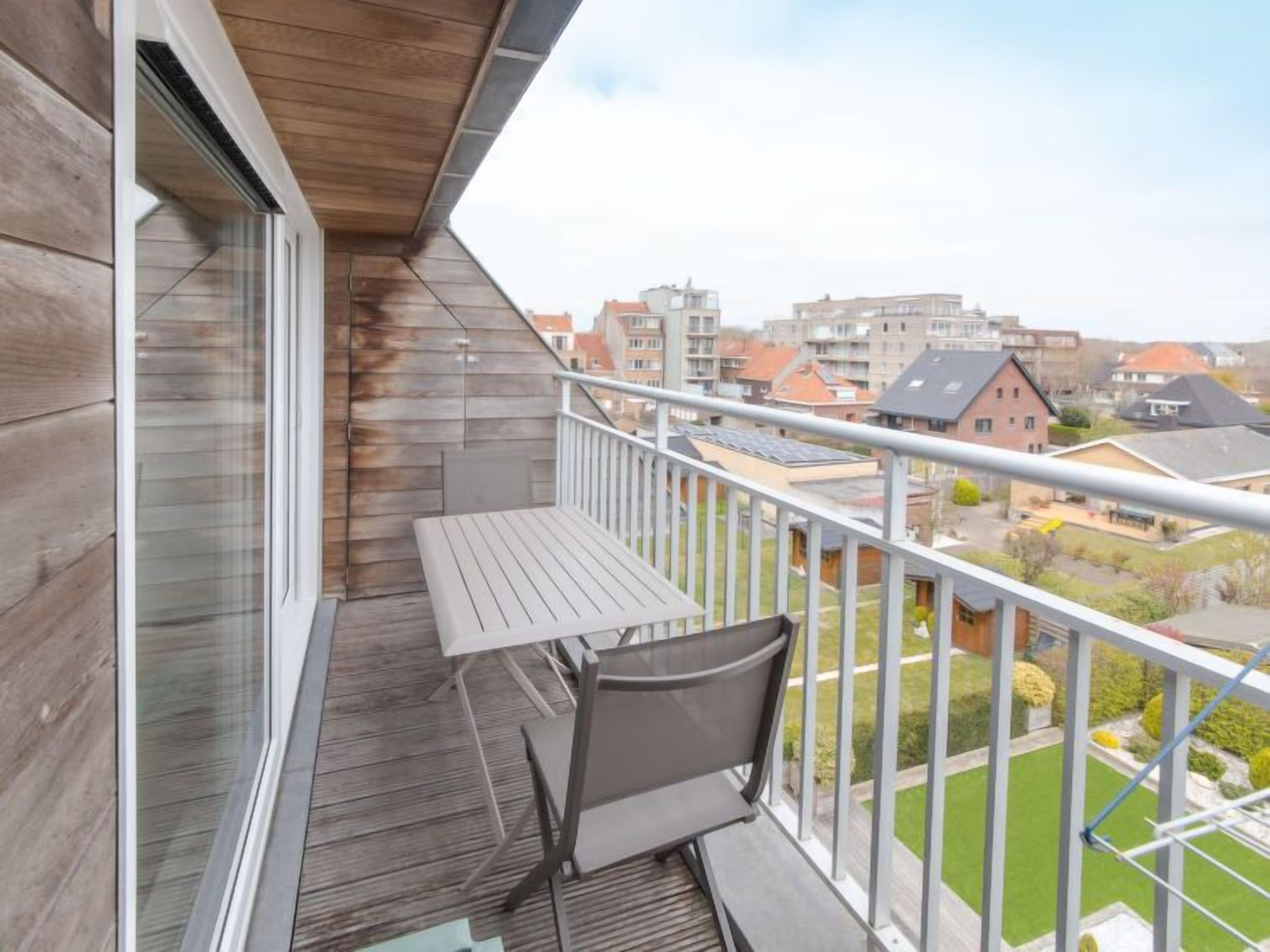 A balcony with a table and chairs, overlooking a residential area with houses and a green lawn.