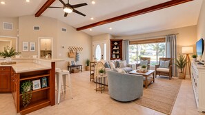 A spacious living room with a sofa, armchairs, and a coffee table. There is a kitchen area with a countertop and stools. The room has large windows with curtains. - Heated Pool + Hot Tub, Game Garage & Teen Shed – 4BR Near Beaches (Bradenton)