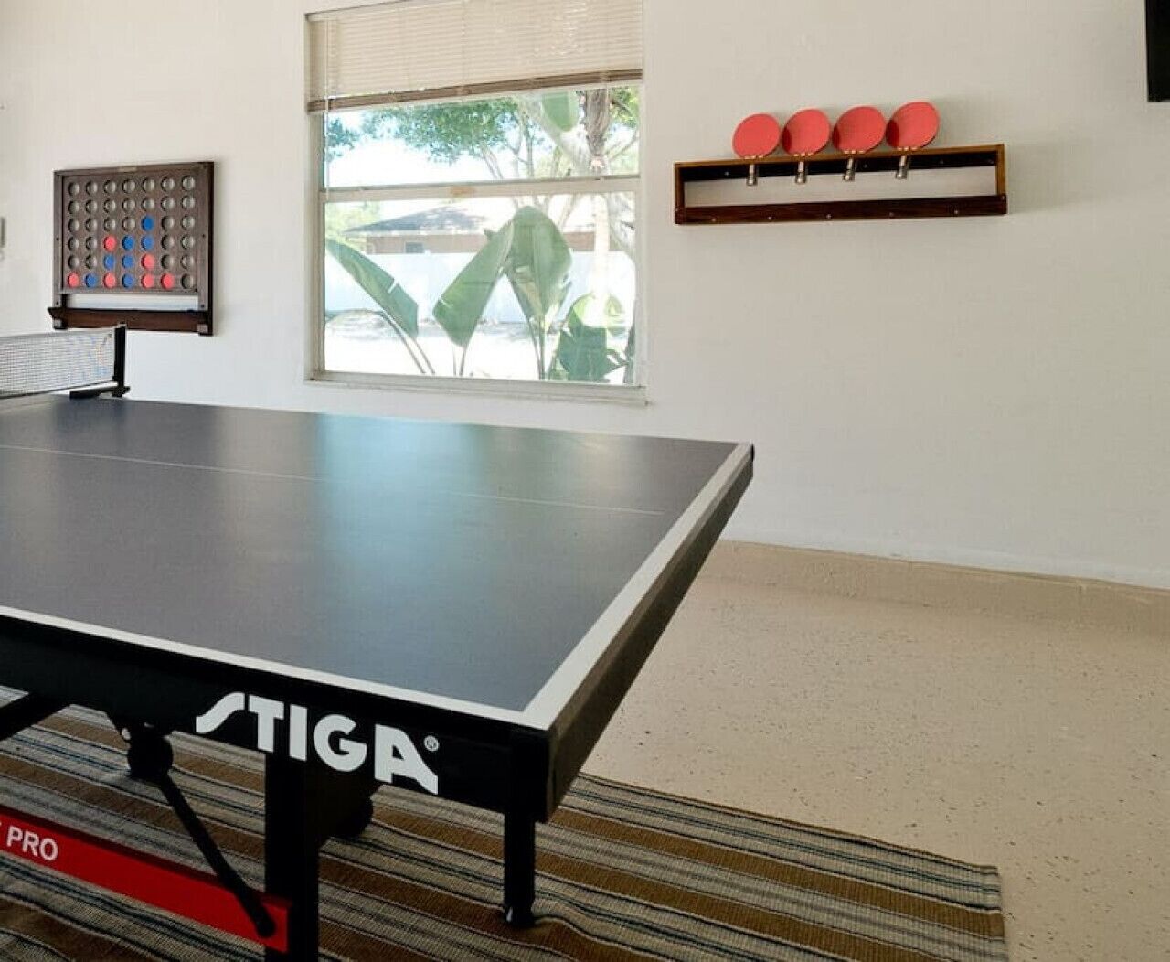 A ping pong table with a net, a wall-mounted table tennis paddle rack, and a window with a view of greenery.