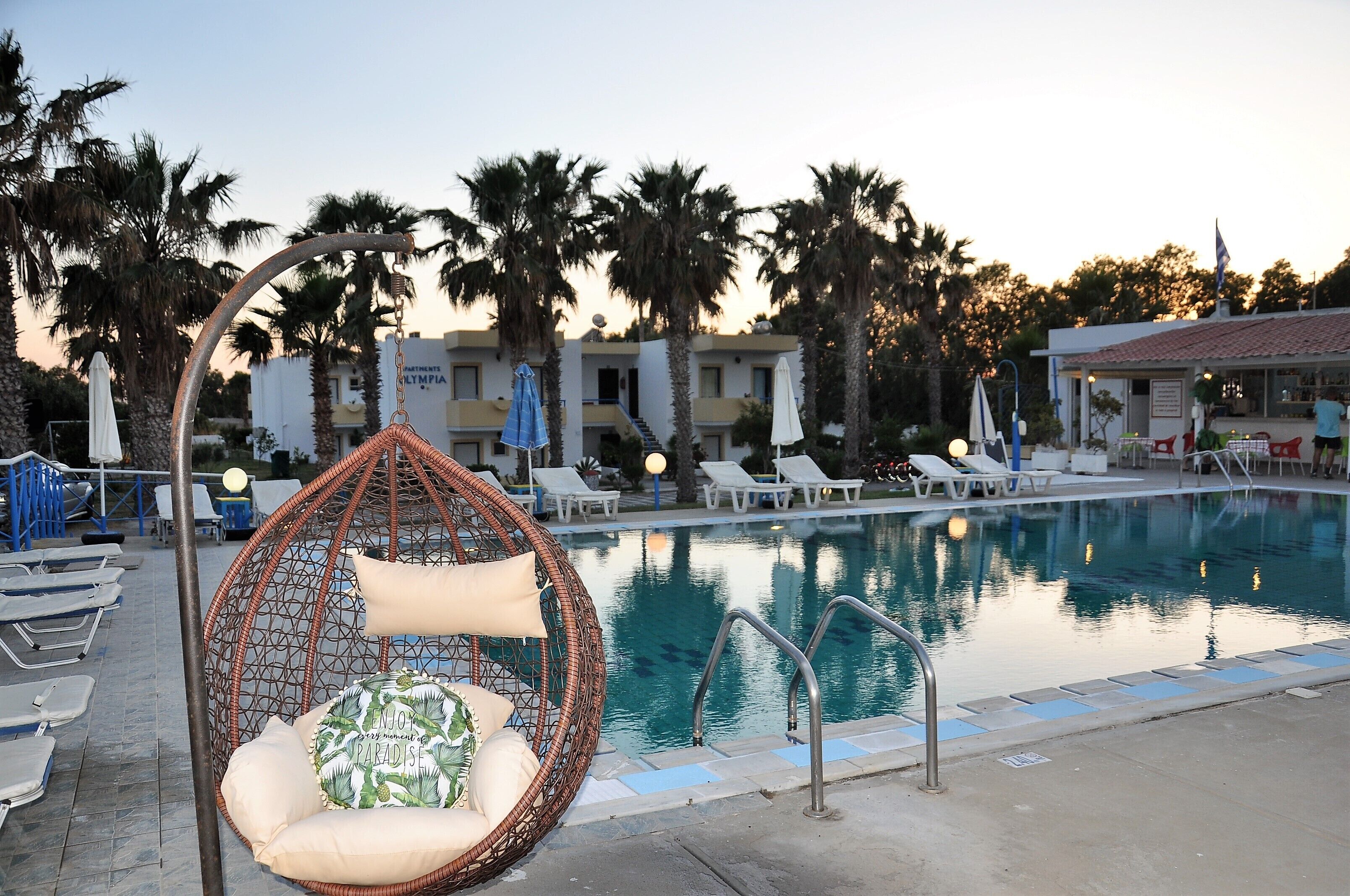 Un espace piscine avec des chaises longues, un fauteuil suspendu et, au fond, un bâtiment portant l’inscription « APARTMENTS LYMPIA ».