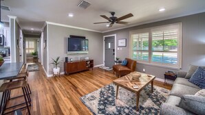 A spacious living room with hardwood floors, a ceiling fan, a large window with blinds, and a TV mounted on the wall.