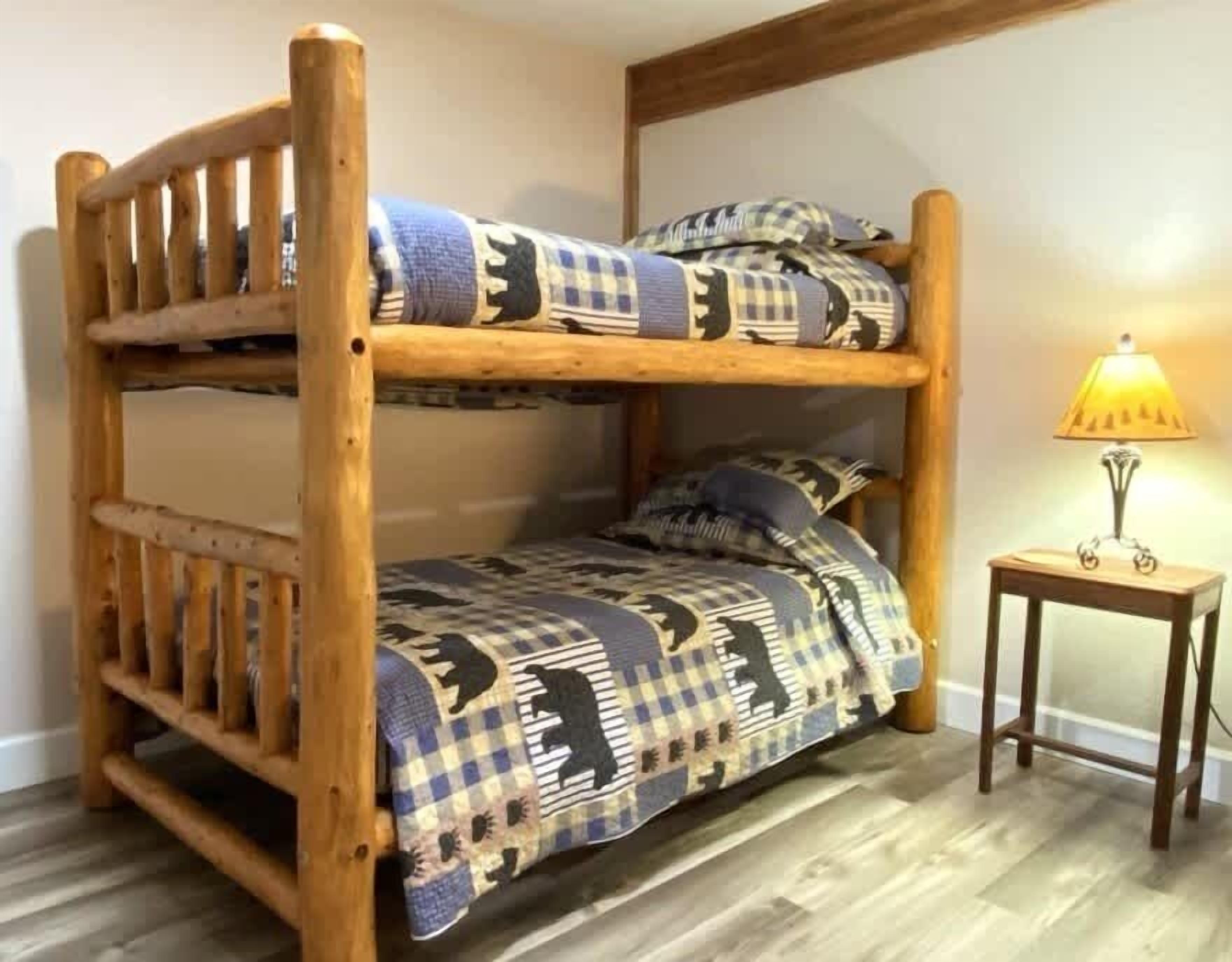 A bunk bed with a checkered blanket and pillows, a small wooden side table with a lamp, and a wooden floor.