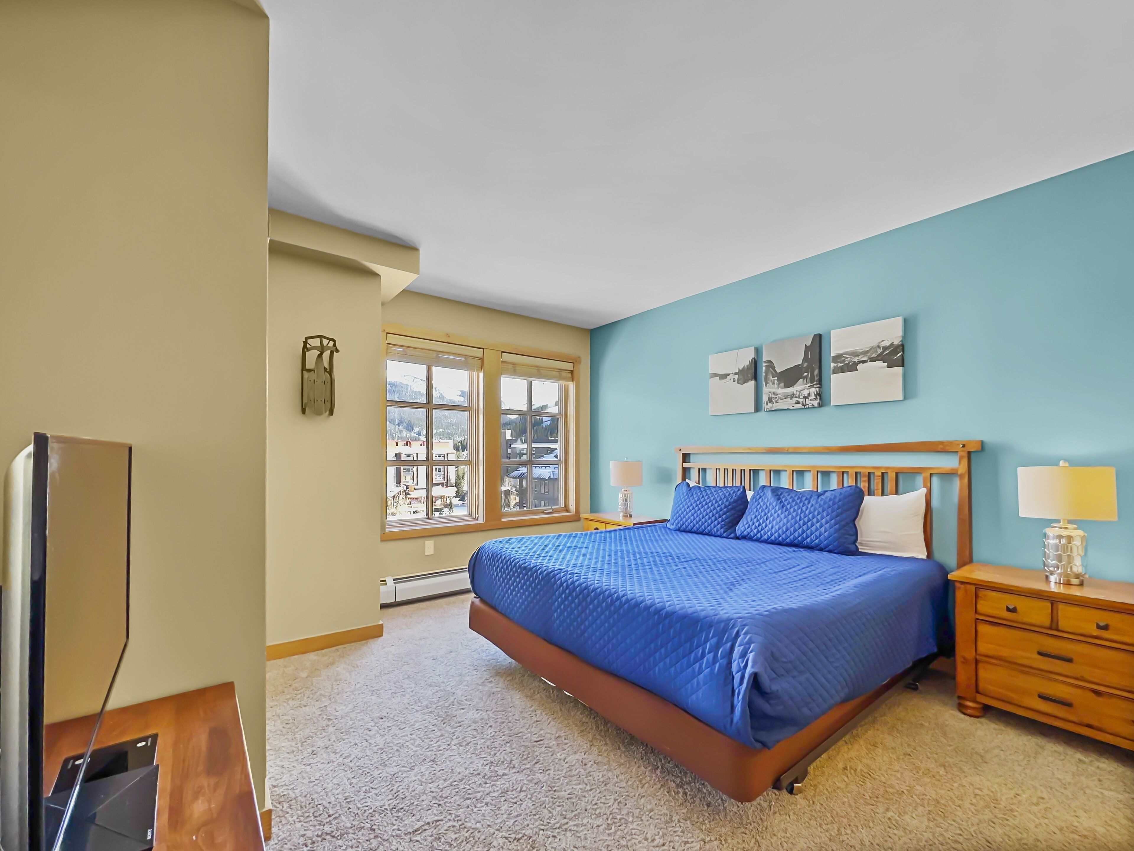 A bedroom with a large bed, a wooden nightstand, a TV, and a window with a view of buildings.