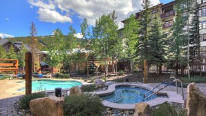 A pool area with a hot tub, surrounded by rocks and greenery, with buildings in the background. - Luxury Remodeled Passage Point Condo - Short Walk to Lifts - Incredible View - PP403 by Redawning (Copper Mountain)