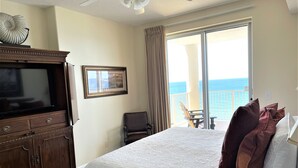 Una camera d'albergo con un grande letto, una televisione, un quadro incorniciato, una sedia e un balcone con vista sull'oceano.