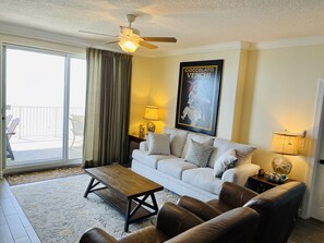 A living room with a sofa, coffee table, and leather chairs. There is a ceiling fan, a painting, and a window with curtains.