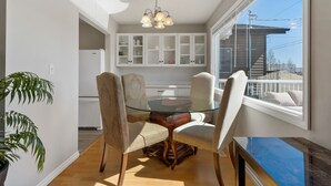 Un coin repas avec une table en verre, quatre chaises et un lustre. Au-dessus de la table, on trouve des meubles blancs et une fenêtre d’où l’on peut voir une maison et des lignes électriques.